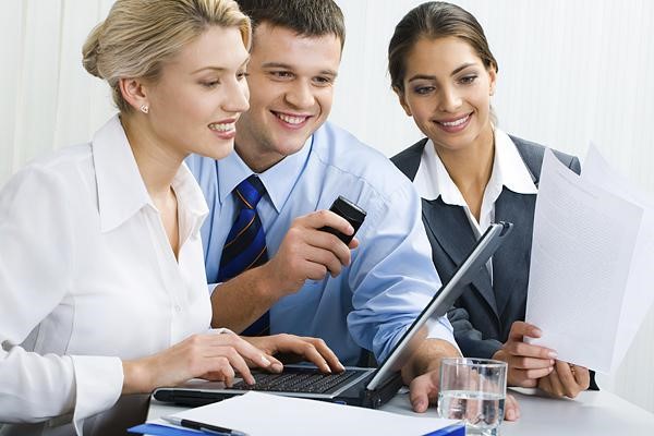 Three people are sitting at a table and one is using his laptop.